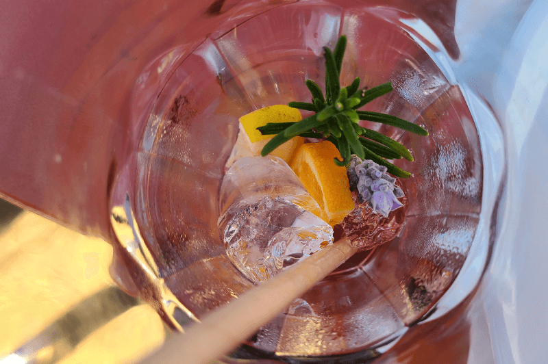 Empty glass of orange blossom iced tea with ice cubes still present, orange, a sprig of rosemary, and a lavender bloom